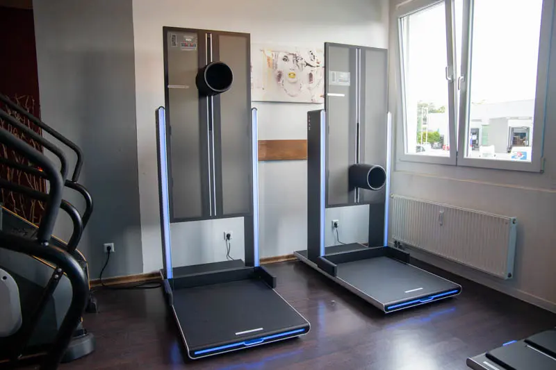 Zwei moderne Fitnessgeräte stehen nebeneinander in einem gut beleuchteten Fitnessstudio in der Nähe eines Fensters. Jedes Gerät verfügt über eine elegante schwarze Plattform mit vertikalen Paneelen, in die Monitore und große runde Aufsätze eingebaut sind. Das Fitnessstudio hat dunkle Holzböden und ein Wandgemälde.